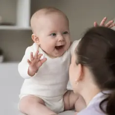 Radosne gaworzenie maluszka do mamy. Dziecko w białym body wyciąga rączki, uśmiechając się szeroko.
