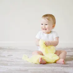 Uśmiechnięte roczne dziecko w białym body bawi się żółtą tkaniną na drewnianej podłodze.