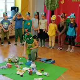 Dzieci w strojach zwierząt i zielony smok uczą się segregacji śmieci. To lekcja o ekologii, która może być inspiracją do krótkich wierszyków.