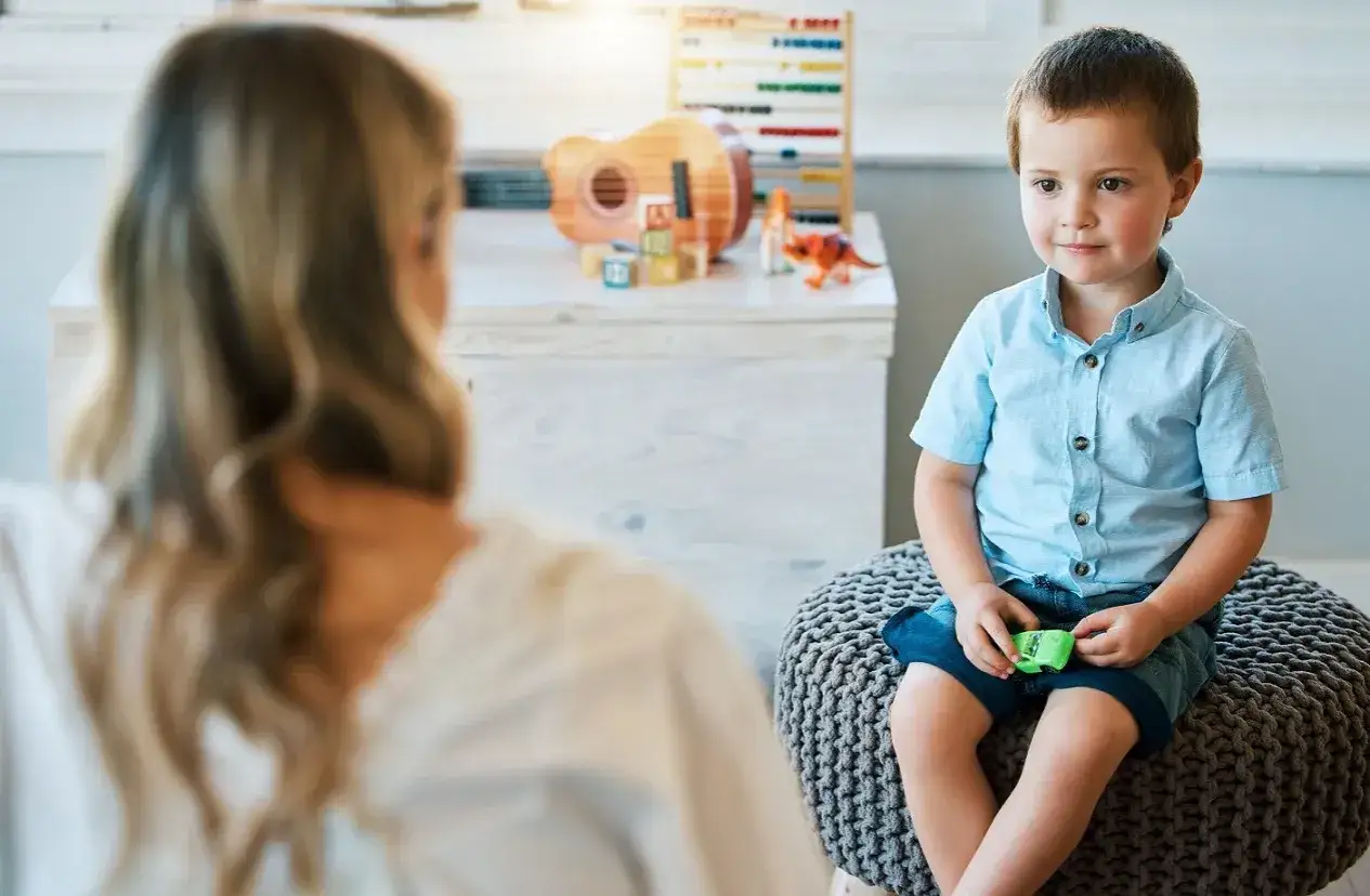 Chłopiec z zespołem Aspergera siedzi na pufie, rozmawiając z terapeutką. W tle zabawki.