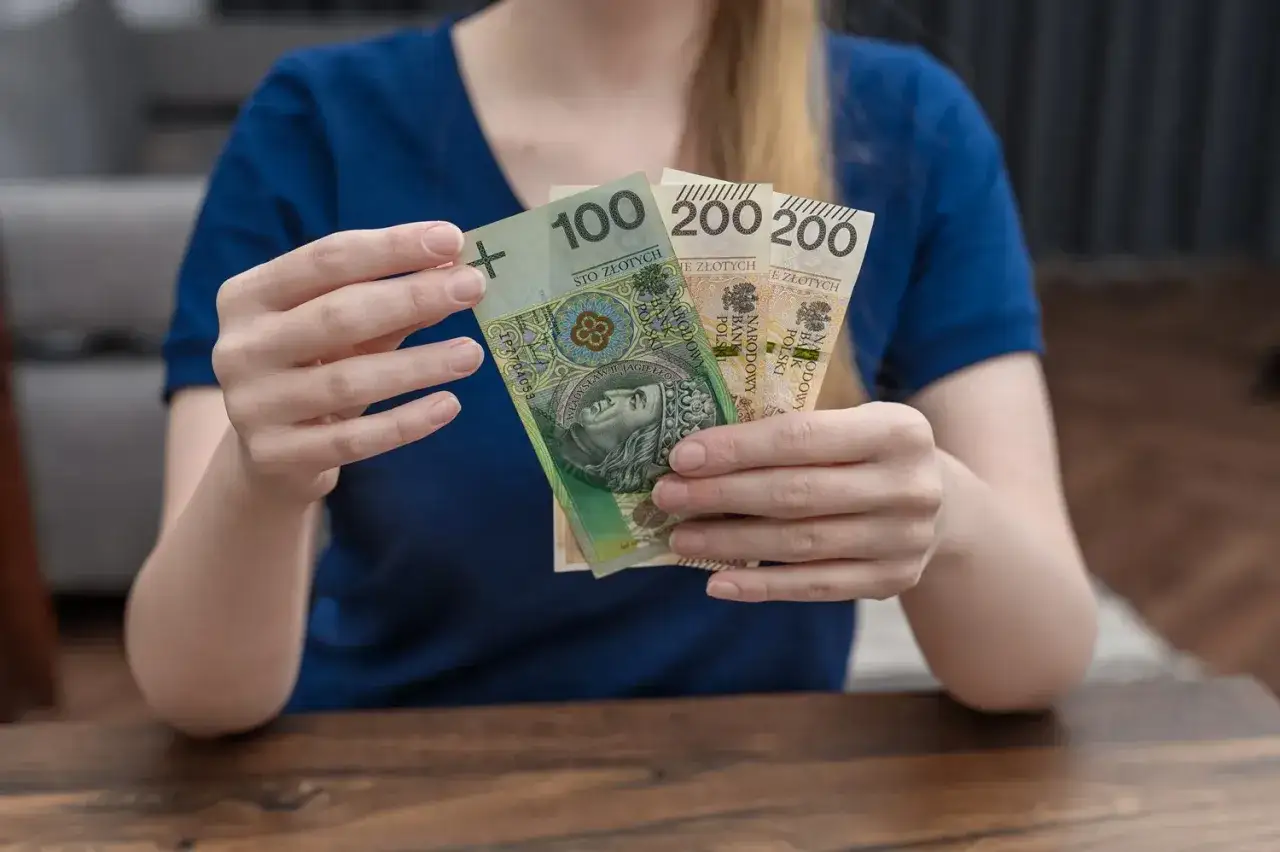 Dłonie trzymają polskie banknoty: 100 zł i dwa po 200 zł. Może to być część zasiłku rodzicielskiego.